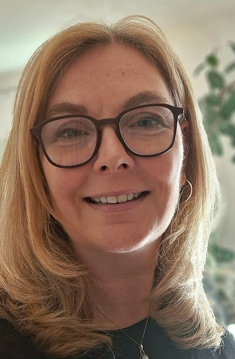 Frau mmit  braunem Pulli und blonden mittellangen Haaren liegt mit den Armen aufgestützt auf der Wiese und schaut dabei lachend in die Kamera.