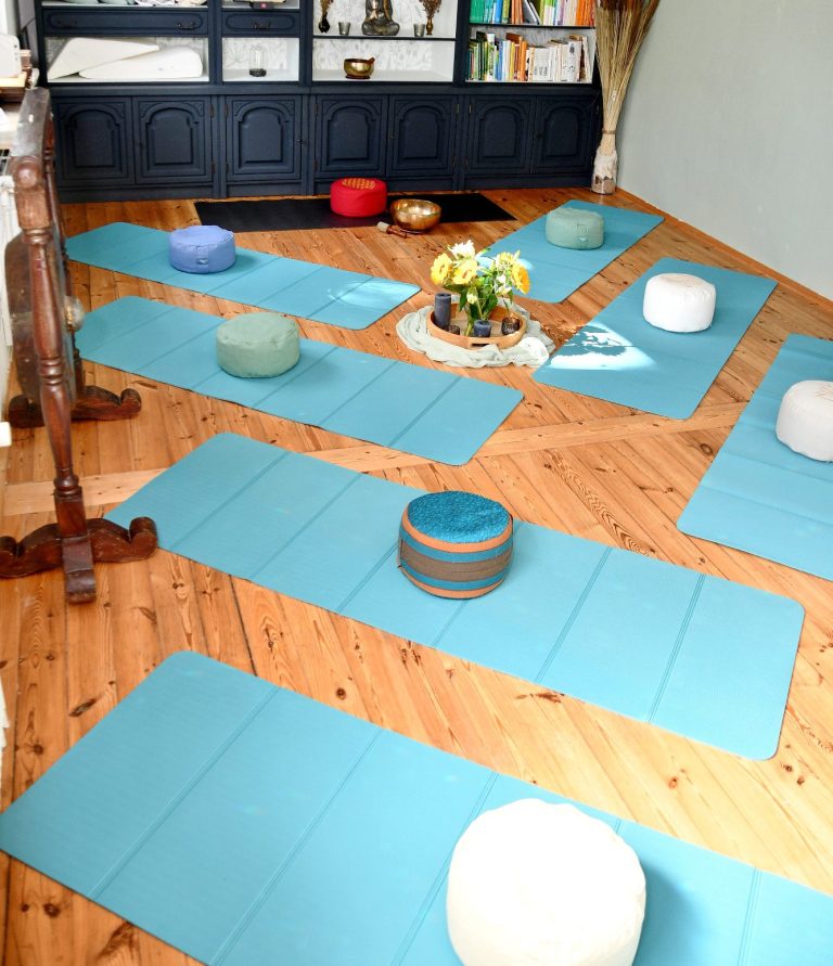 Sechs türkisfarbene Bodenmatten mit Meditationkissen darauf auf einem Holzfußboden. Ein Tablett mit Blumen und Kerzen dazwischen. Am Ende eine Klangschale auf einer dunklen Matte mit einem roten Kissen.