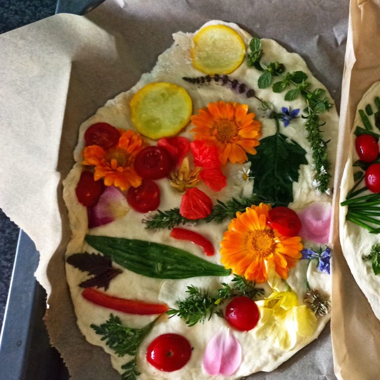 Ein flacher Teigfladen auf einem Blech. Der Falden ist dekoriert mit Blüten, Blättern, Tomaten und Zucchini. Ein essbares Blumenbild. 