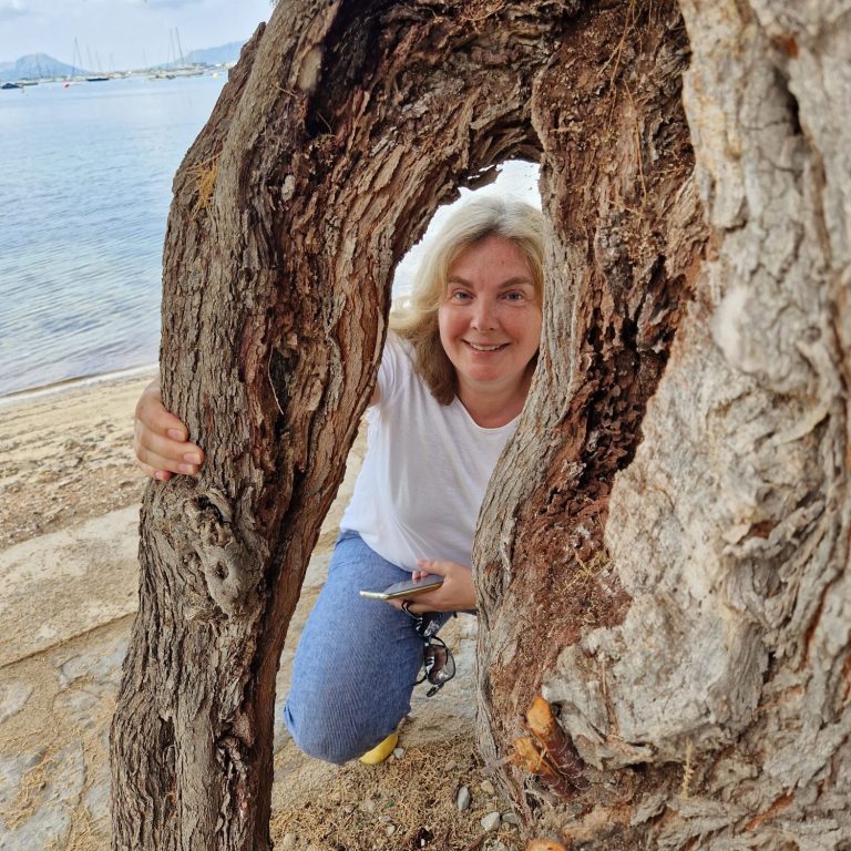 Frau mit Jeans und weißem T-shirt schaut fröhlich zwischen den Wurzeln eines sehr alten und großen Baumes hindruch. Im Hintergrund das Meer.