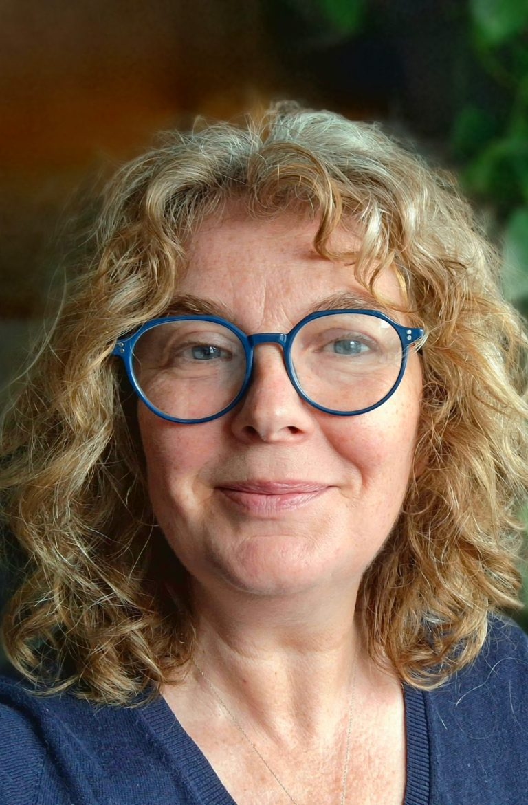Frau mmit  braunem Pulli und blonden mittellangen Haaren liegt mit den Armen aufgestützt auf der Wiese und schaut dabei lachend in die Kamera.