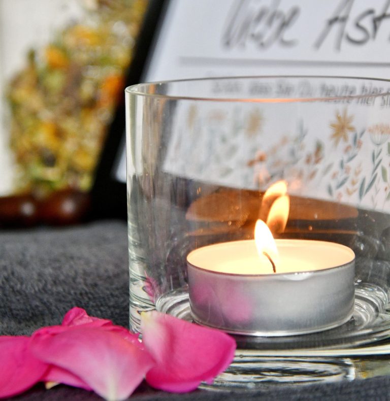 Eine Glas mit einem brennenden Teelicht steht auf einem grauen Handtuch. Darauf sind Rosenblätter in Rosa. Dahinter ein Willkommens-Schild mit schwarzem Rahmen und Blumendeko.